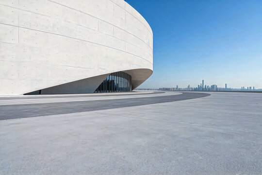 This shot shows a curved modern concrete building exterior with an empty open plaza, under a clear blue sky with distant city skyline.