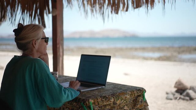 Mature woman focuses on work at her laptop while enjoying the beach and ocean view in Baja California Mexico.