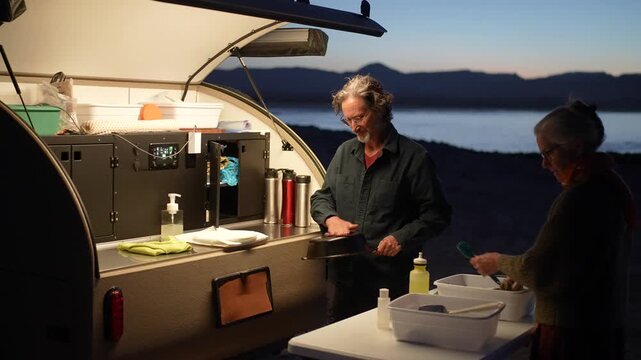 A mature woman cleans dishes by a teardrop camper while enjoying the sunset at the beach in Baja California, Mexico.