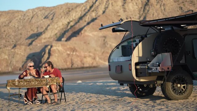 A mature man and woman dine at a table near their teardrop camper trailer on the beach in Baja California during sunset.