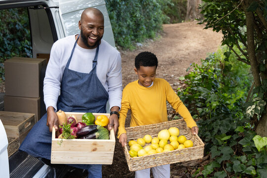 African American father and son laughing, in apron holding crate, son holding lemons at white van