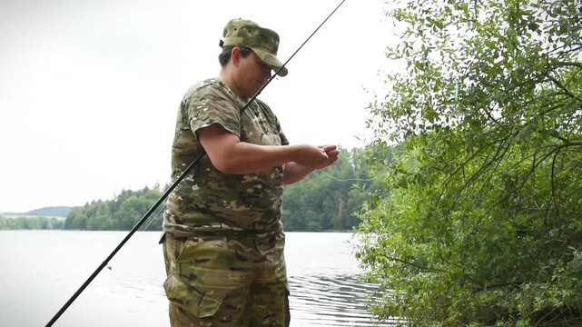 Woman in camouflage clothing prepares her fishing rod and bait on the shore of a lake