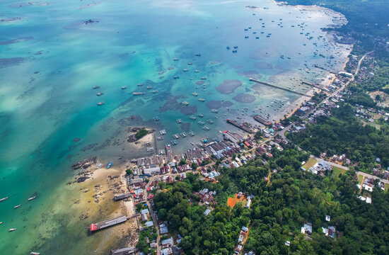High-angle aerial view of a traditional fishing village in Tanjung Binga, Belitung, Indonesia, featuring coastal houses, fish drying platforms, and boats on turquoise water.