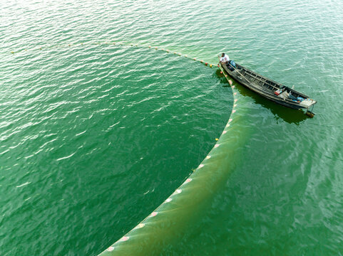 Aerial view of a fisherman in a wooden boat pulling a large green net in the calm green waters in Rangamati, Chittagong Division, Bangladesh.