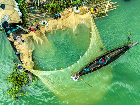 Aerial view of fishermen on wooden boats pulling a large yellow fishing net in the green water in Rangamati, Chittagong Division, Bangladesh.