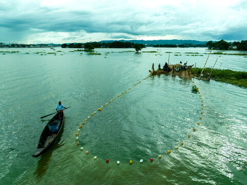 Aerial view of fishermen in wooden boats casting a large circular fishing net with colorful floats on the calm waters of Kaptai Lake under an overcast sky Rangamati, Chittagong Division, Bangladesh.