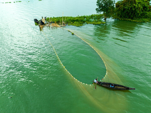 Aerial view of fishermen in wooden boats casting a large circular net in the green waters of Kaptai Lake near a small lush island in Rangamati, Chittagong Division, Bangladesh.