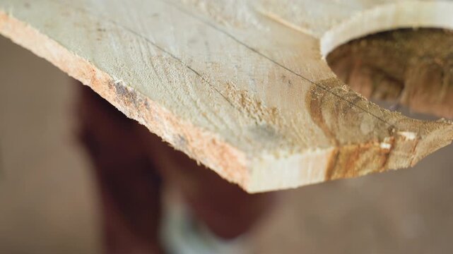 Angled view curved wood edge with circular cutout, rough fibers and sawdust along perimeter, blurred carpenter in background, warm workshop atmosphere, board being inspected for fit and final