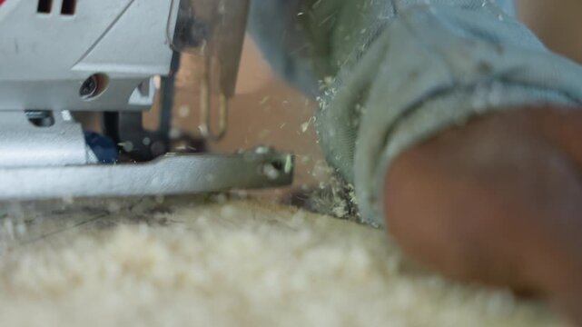 Closeup jigsaw guided by hand, skilled carpenter guiding blade across plank, sawdust spray and wood chips, visible grip and forearm pressure, intimate workshop scene showing craft and technique