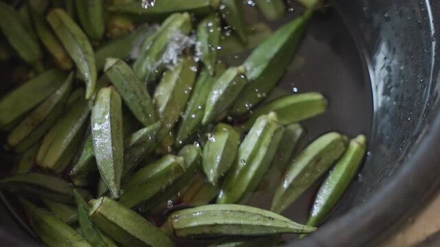 Fresh okra pods rinsing in bowl, closeup view of green pods floating in shallow water with gentle pour, subtle droplets on skin, rustic black bowl under natural kitchen light, food prep sequence