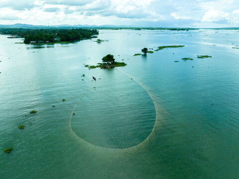 Aerial view of Kaptai Lake featuring a large fishing net spread in the water near a small island with a lone tree and a traditional boat Rangamati, Chittagong Division, Bangladesh.