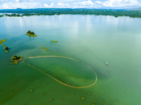 Aerial view of circular fishing nets in Kaptai Lake with small islands and a boat under a bright sky in Rangamati, Chittagong Division, Bangladesh.