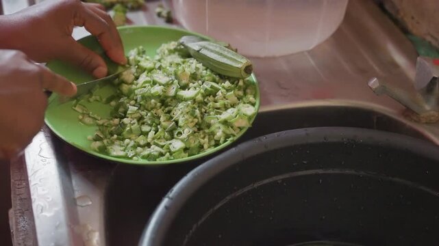 Closeup black hands chopping okra on green plate over sink, methodical cutting motion, fine pieces falling into bowl, warm kitchen light, domestic meal prep, textured skin and fingertip detail showing