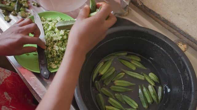Sink hands sorting okra into pot, marketstyle prep with chopped greens on plate, large knife, wet fingertips, fastpaced communal cooking, busy countertop, closeup of motion and texture, vibrant