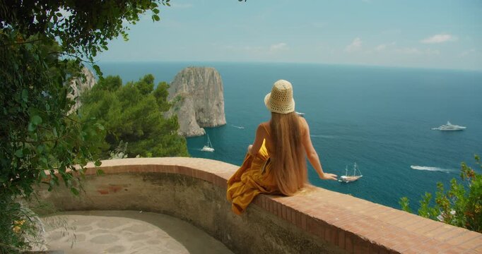 Woman in summer dress enjoying seascape while sitting on Capri terrace and admiring Faraglioni sea cliffs