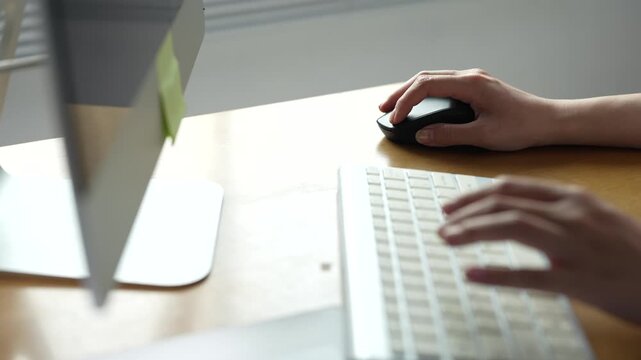 Professional woman using a computer for online work, typing on the keyboard and clicking the mouse on a desk with a monitor and a post-it in an office setting