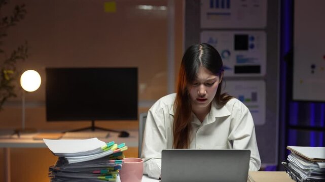 Stressed korean businesswoman experiencing a nervous breakdown from overworking late at night at her office desk with a laptop and a large pile of paperwork