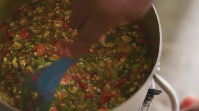 Topdown pot stirring vibrant vegetable relish with green spoon diced peppers, tomatoes and okra mixed over medium heat, hands guiding spoon, tutorial style pacing, sizzling sound and aromatic steam,