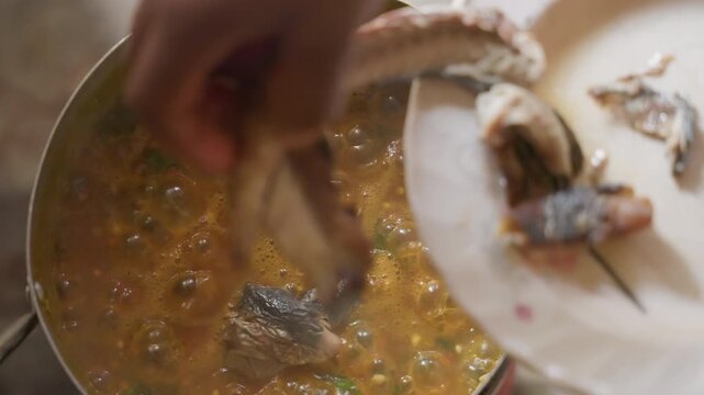 Closeup simmering fish stew in pot, hand drops whole fish into bubbling spiced broth, steam rising, visible bones and herbs, intimate rustic kitchen scene, slow attentive motion by home cook, earthy