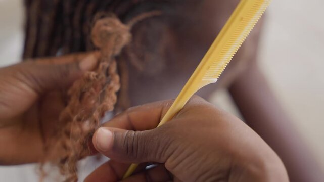 Closeup stylist combing braid ends with applicator and tube, careful squeeze and comb motion shapes extension tips, focused hands show dexterity and finishing technique under soft studio light, salon