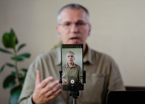 Mature man recording an educational video or webinar on a smartphone. Focus on the phone screen showing the speaker gesturing. Professional vlogging and remote teaching concept.