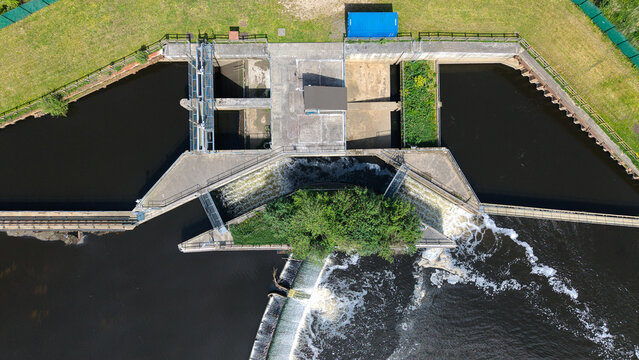 Aerial view of Knottingley Weir and Hydroelectric Power Plant with water rushing over the concrete spillway and green riverbanks in Knottingley, England, United Kingdom.