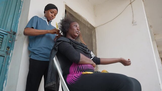 Black trainee styling seated client confidently, stair landing or balcony setting, young stylist finishing curl arrangement, client smiling with relaxed posture, supportive mentor presence, candid