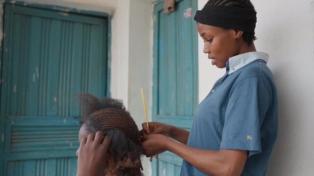Black hairstylist braiding hair at door, woman in blue shirt gently sections clients afro with yellow comb, focused expression, warm natural light, intimate neighborhood setting, calm caring