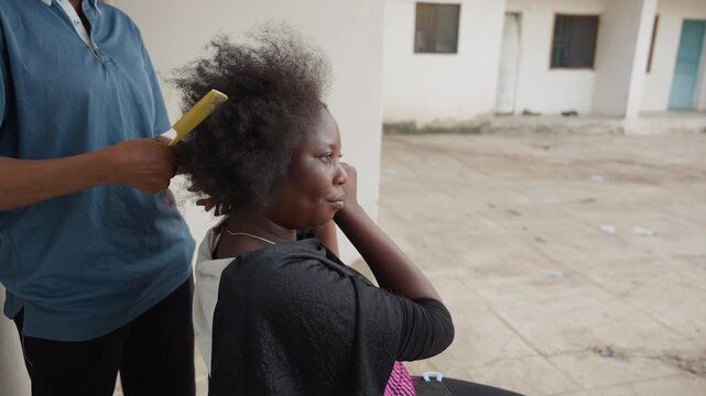 Elderly black woman having hair combed outside, young assistant in blue shirt, gentle hands styling gray hair, courtyard background, relaxed posture, intimate caregiving scene, candid documentary