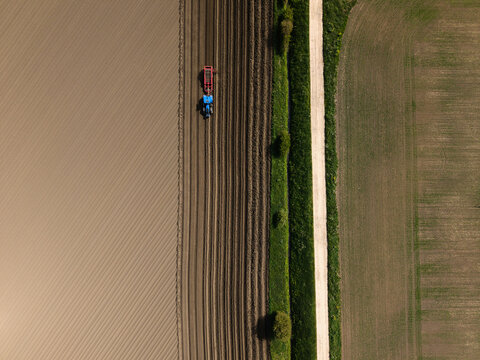 Aerial view of a blue tractor pulling a red implement in a brown agricultural field with parallel tilled lines next to a green grassy path in Knottingley, England, United Kingdom.