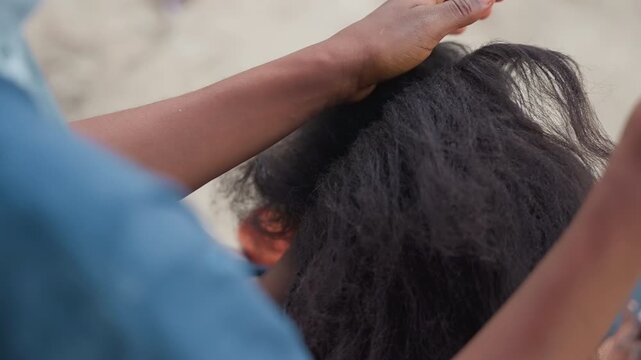 Closeup combing black hair outdoors, focused stylist using metal pick to section and detangle, rhythmic hands weaving braid, urban bench setting, precise parting and neat technique, close attention