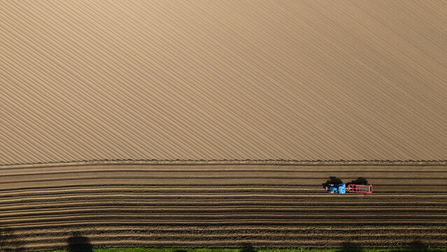 Aerial view of a blue tractor with a red trailer tilling a large agricultural field with distinct diagonal furrows and brown soil Knottingley, England, United Kingdom.