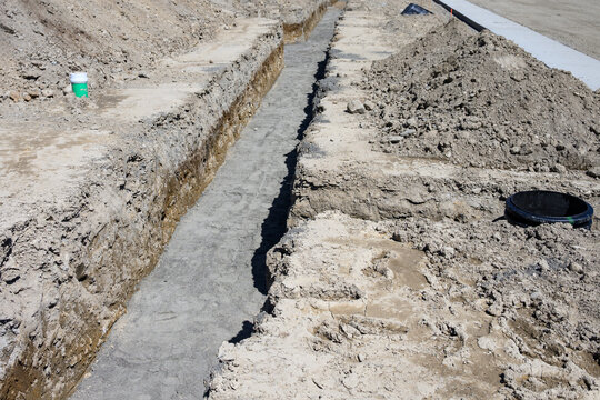 Earthworks in progress on large housing development site, trenches dug for plumbing and water systems, including septic tank riser, road graded and concrete sidewalk poured
