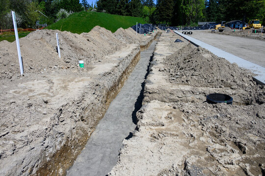Earthworks in progress on large housing development site, trenches dug for plumbing and water systems, including septic tank riser, road graded and concrete sidewalk poured
