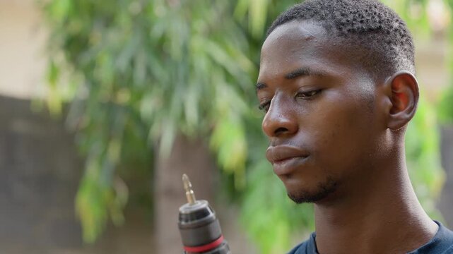 Black handyman testing drill bit closeup, outdoor leafy scene, concentrated expression and sweat, adjusting chuck and checking rotation, preparing for precision woodworking and renovation task,