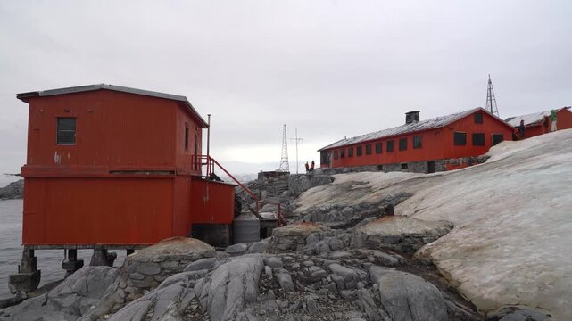 A wide stationary shot of the bright orange research buildings at Argentina's Melchior Base, situated on the rugged, rocky landscape of Observatory Island in Antarctica