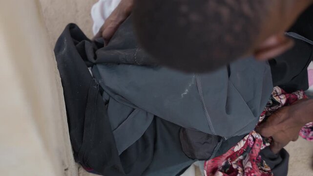Person sorting piled clothing on step, topdown view of hands and layered garments, careful selection and folding of worn fabrics, patched textures and muted tones, quiet concentration