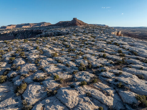 Aerial view of expansive slickrock plateaus with scattered desert vegetation and a prominent rugged butte under a clear blue sky.
