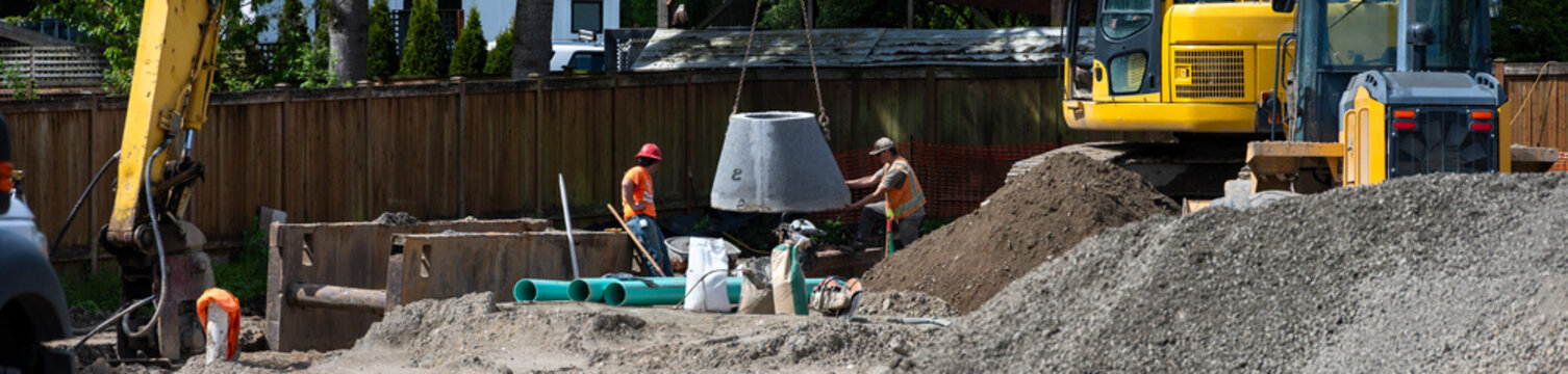 Excavator boom arm being used to lower precast concrete stormwater system access vault into trench, construction workers guiding the installation process
