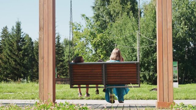 Child and parent on swing under bright summer trees, blue dress and swinging legs, joyful pointing gesture and laughter, sunlight on grass and carefree weekend energy capturing imaginative outdoor