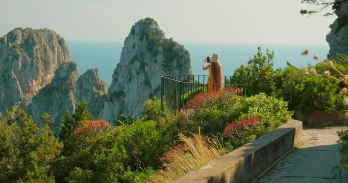 Woman in summer dress recording video of Faraglioni sea stacks in Capri from garden overlook at golden hour