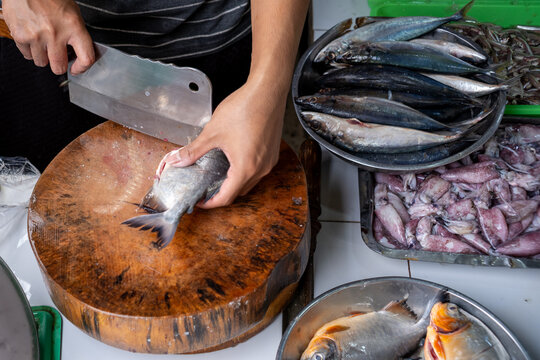 Close-up of a vendor cleaning fresh pomfret (ikan bawal) with a knife on a wooden chopping block, with squid and mackerel displayed at a traditional seafood market in Jakarta.