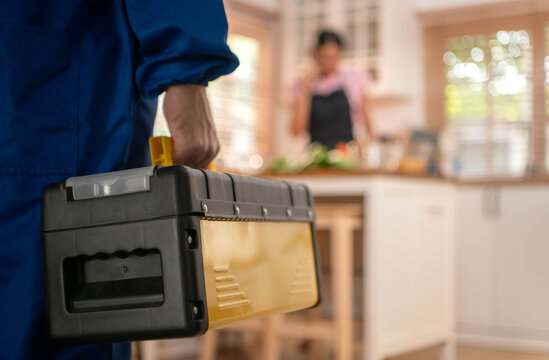 Woman cooking in kitchen while talking on mobile phone as technician arrives with toolbox