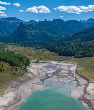 Aerial view of a turquoise mountain lake with low water levels revealing sandy banks and braided river channels surrounded by lush green forests and jagged peaks.
