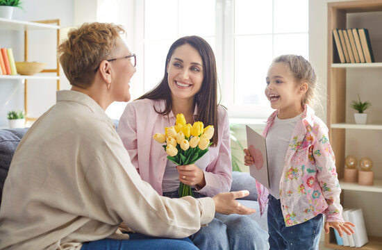 Happy cheerful little child girl with mother congratulating senior mature grandmother on holiday with greeting card and flowers bouquet sitting on sofa at home. Family and mothers day concept.