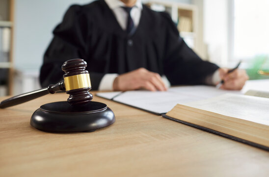 Judge's gavel on workplace with male judge working in background. Male justice worker wearing judicial robe with mallet in courtroom having court verdict. Jurisdiction and legal law concept.