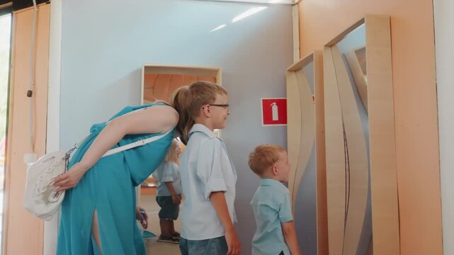 Mother and boys testing warped mirror, woman in turquoise dress bending over two children inspecting wavy reflection panels indoors, bright interior light, playful laughter and surprise, casual