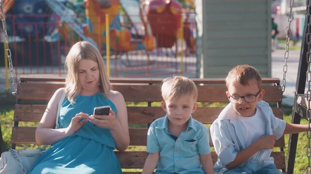 White mother scrolling phone beside boys on bench at park ride during weekend, children fidgeting and glancing at surroundings, warm sunlight and carnival backdrop highlight modern parenting contrast