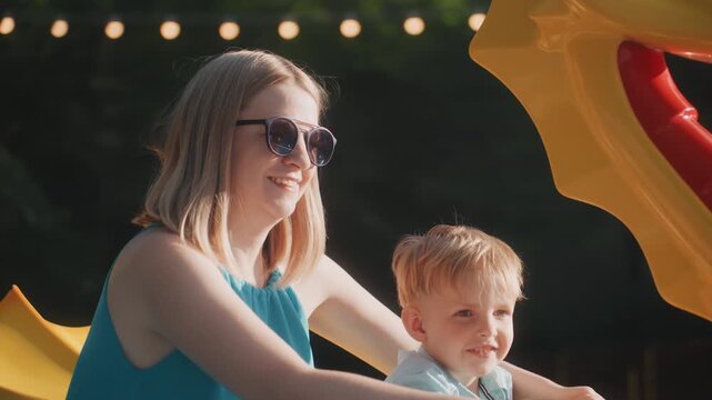 Caucasian mother with son on ride, dragon carousel framed with string lights, smiling toddler and parent bonding, joyful carnival energy, playful weekend family outing, caregiver guiding, candid
