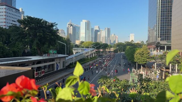 Wide horizontal view of a large crowd during car-free day along Jalan Jenderal Sudirman with modern skyscrapers in the background and soft blurred flowers in the foreground. People walking, jogging.